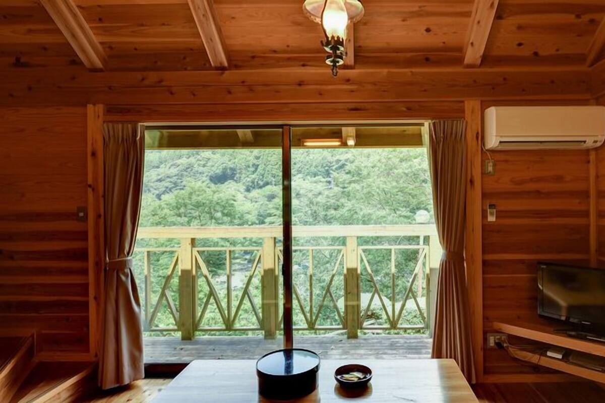 Une cabane en bois avec une table à manger, un bol, un téléviseur et une vue sur une forêt, visible à travers une porte coulissante.
