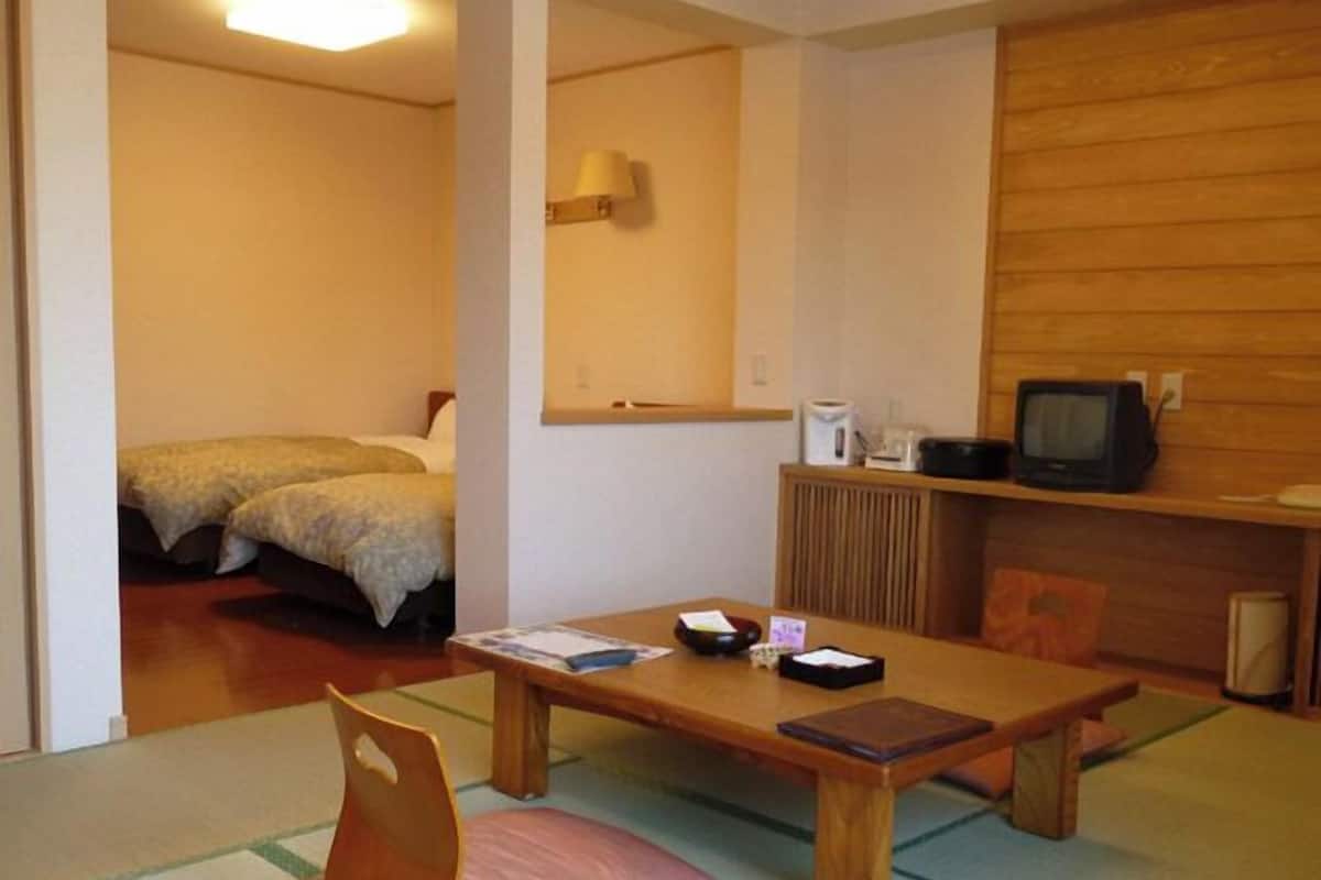 Ein Zimmer mit einem Holztisch, einem Stuhl, einem Bett, einem Fernseher und einem Regal mit Wasserkocher und Fernbedienung.
