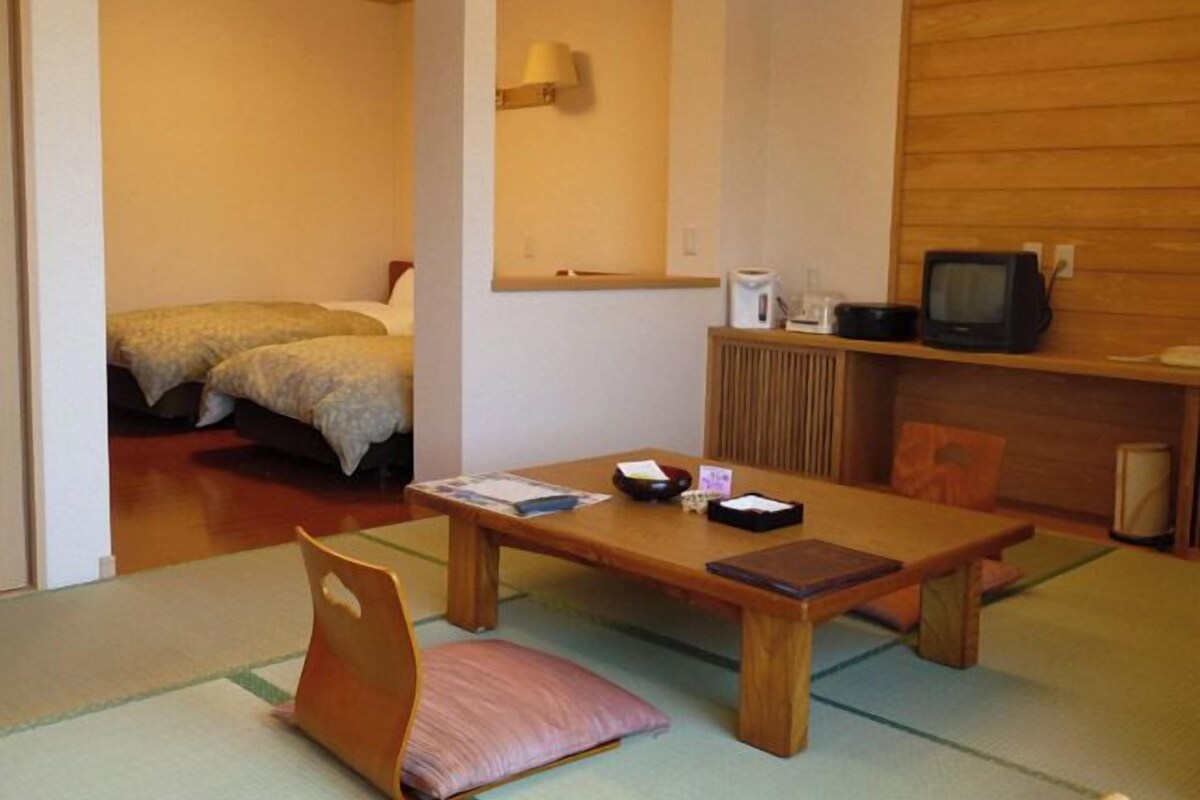 A room with a wooden table, a chair, a bed, a television, and a shelf with a kettle and a remote control.
