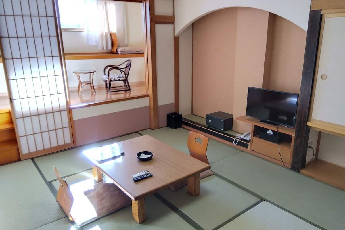 Ein traditioneller japanischer Raum mit Tatami-Boden, einem niedrigen Tisch, Stühlen, einem Fernseher und einer Schiebetür.