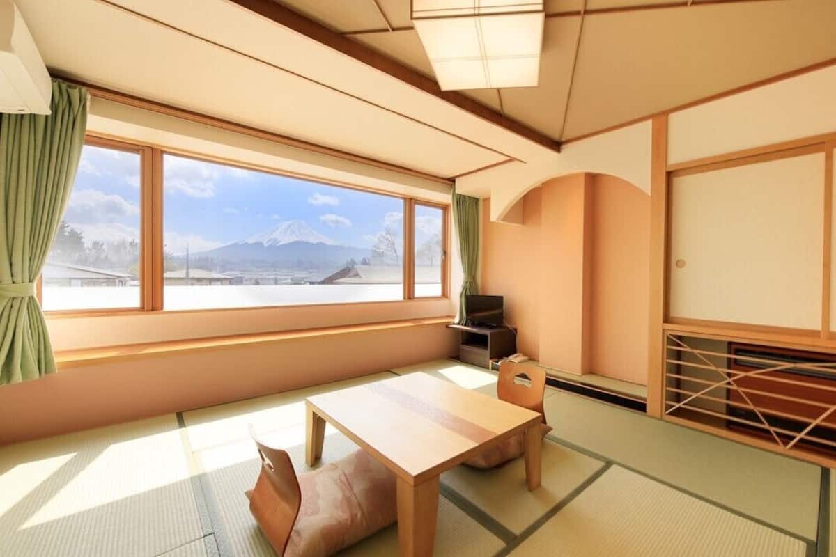 Ein traditioneller japanischer Raum mit Tatami-Boden, einem niedrigen Tisch, einer gepolsterten Sitzbank, einem Fernseher und Blick auf einen Berg.