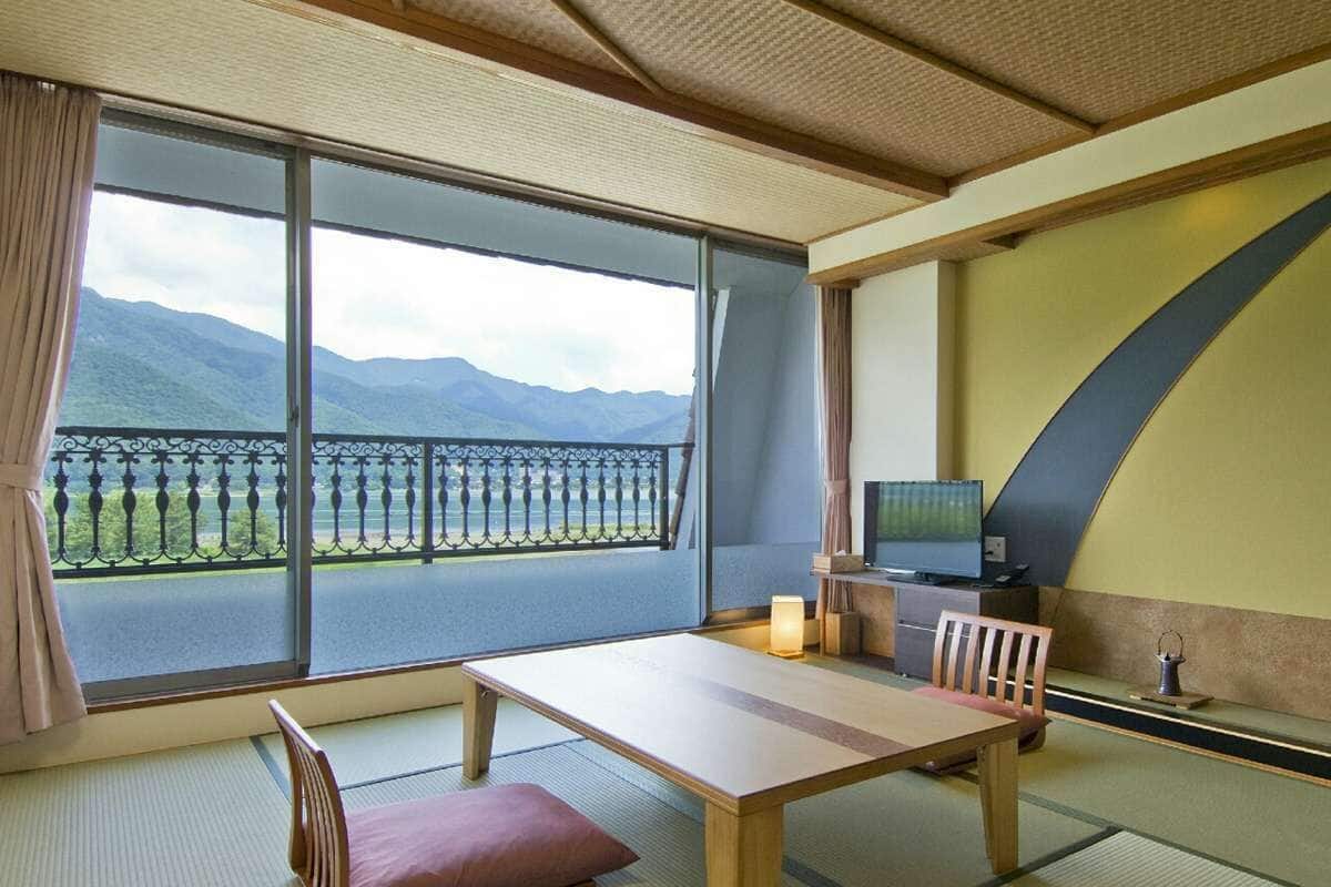 Ein Zimmer mit Tatami-Boden, einem niedrigen Tisch mit Kissen, einem Fernseher und Bergblick durch große Fenster.