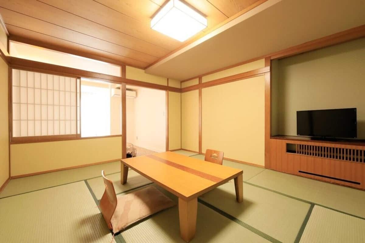 Ein traditioneller japanischer Raum mit Tatami-Bodenbelag, einem niedrigen Holztisch, einem Fernseher und verschiebbaren Türen.