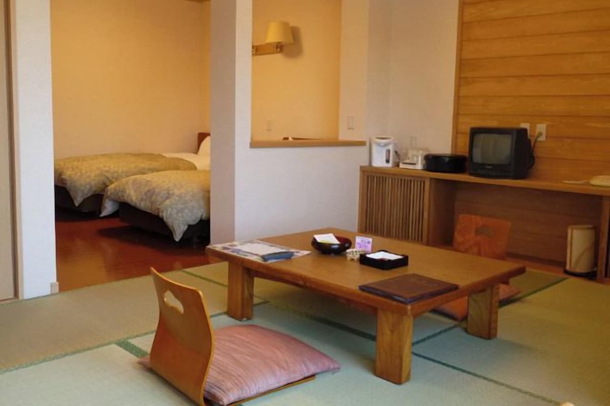 A room with a wooden table, a chair, a bed, a television, and a shelf with a kettle and a remote control.