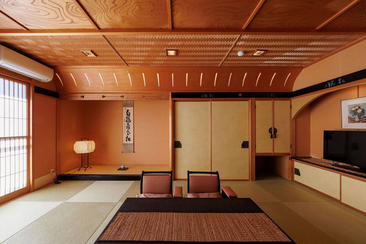 Ein traditioneller japanischer Raum mit Tatami-Böden, einem niedrigen Tisch mit Kissen, einer Tatami-Matte in der Mitte, einer Schiebetür, einem Fernseher und einem Wandbild.
