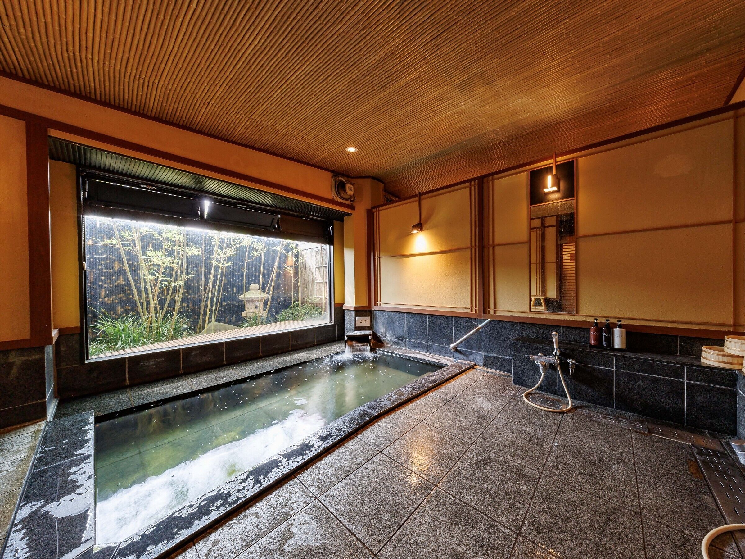 Ein traditionelles japanisches Onsen mit Blick auf Bambus vor dem Fenster, einer Steinsitzbadewanne und einem gefliesten Boden.