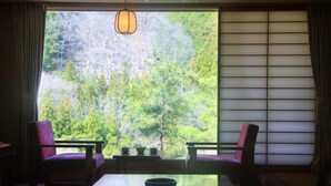 Ett traditionellt japanskt rum med tatamimattor på golvet, ett lågt bord, två stolar och utsikt över en skog genom skjutbara, genomskinliga dörrar (shoji).