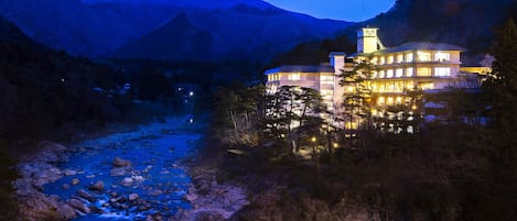 Un hotel con varios pisos, una torre destacada y vistas a un río y montañas al fondo.