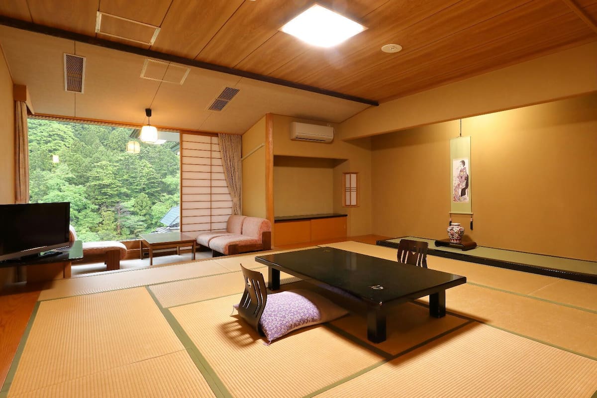 Une pièce avec un sol recouvert de tatamis, comprenant une table en tatami, un canapé, un téléviseur et une porte coulissante donnant sur un paysage verdoyant.