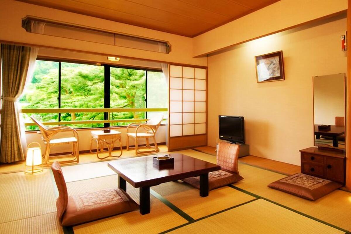 A traditional Japanese-style room with tatami flooring, a low table, a sofa, a television, and a view of greenery outside.