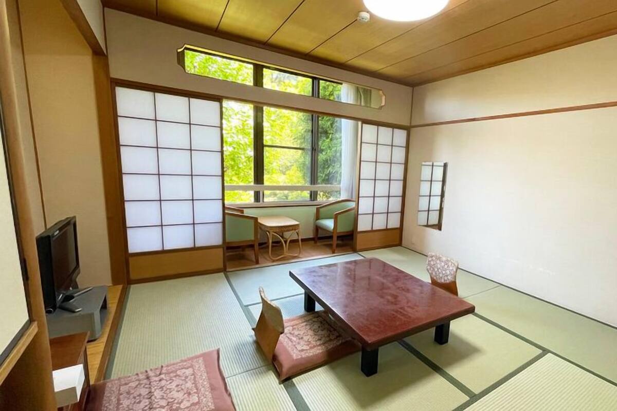 A tatami mat room with a low table, chairs, a television, and a view of greenery outside.