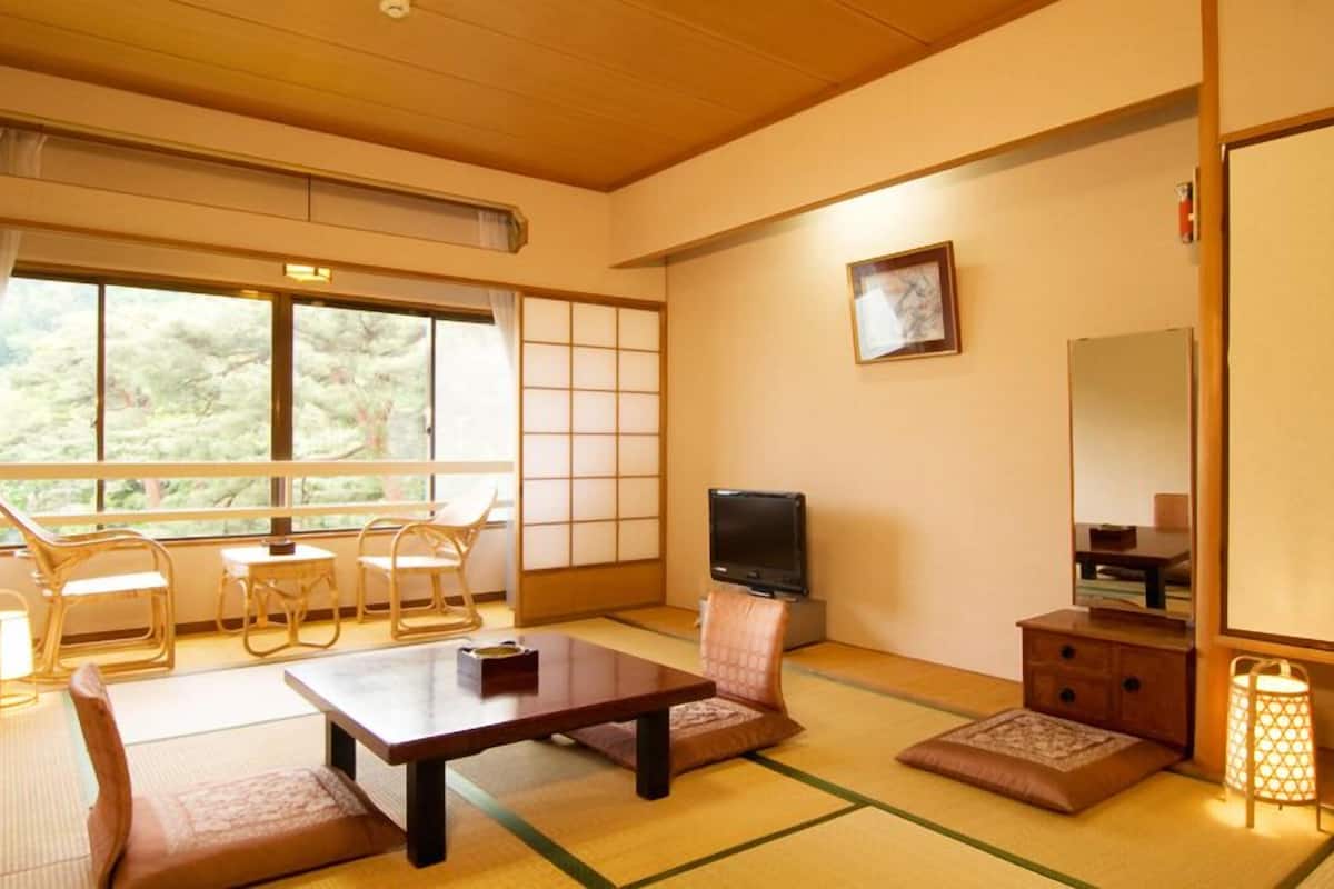 A traditional Japanese-style room with tatami flooring, a television, a sofa, a table, and a view of trees through large windows.