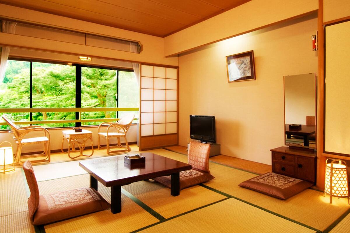 A traditional Japanese-style room with tatami flooring, a television, a sofa, a table, and a view of greenery outside.