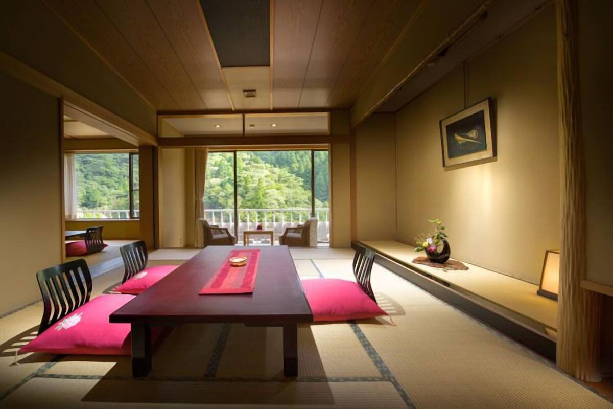 Una habitación tradicional japonesa con suelo de tatami, una mesa baja y vistas a un bosque a través de amplios ventanales.