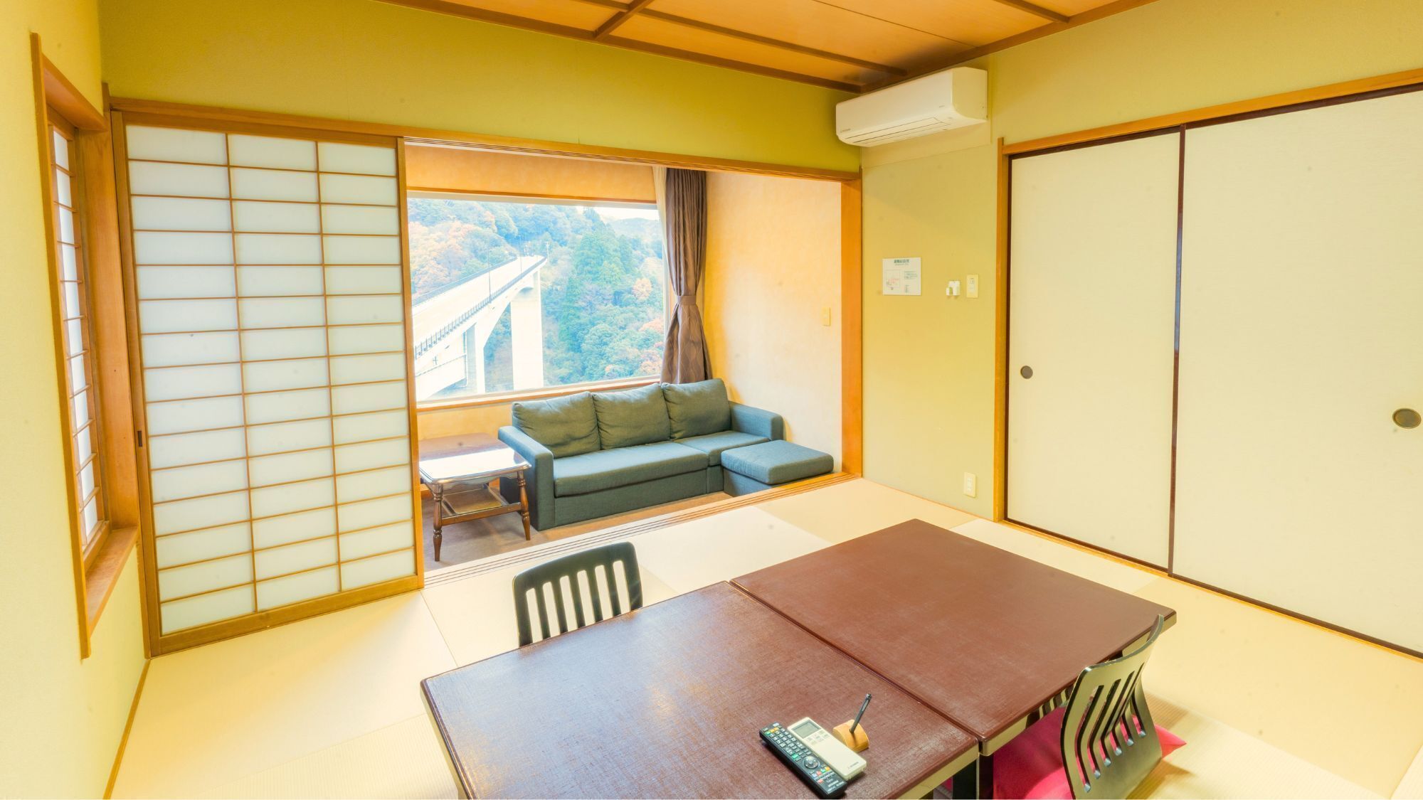A room with a sliding door, a sofa, a table with chairs, and a view of a bridge and trees.