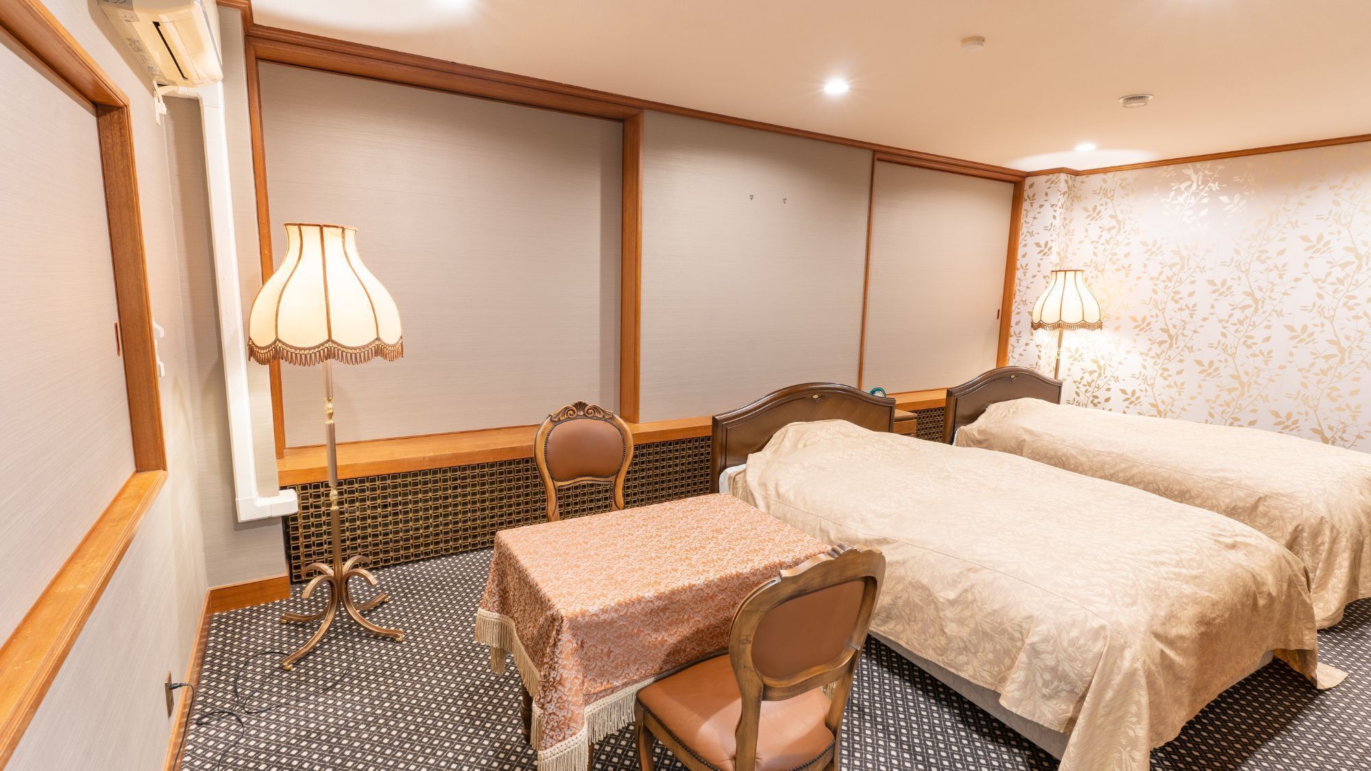 A hotel room with two beds, a table, chairs, a lamp, and a window with blinds.