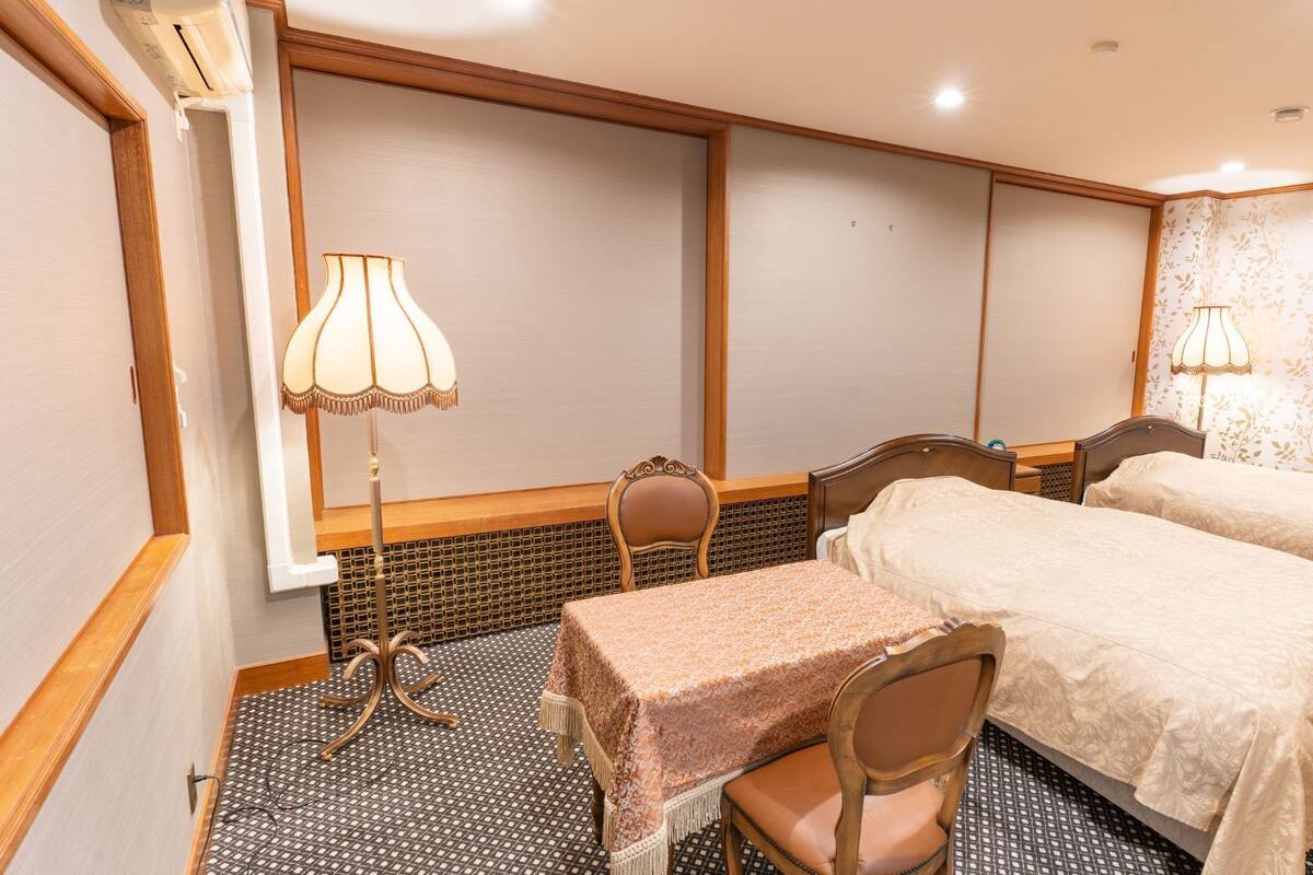 Habitación de hotel con dos camas, una mesa, sillas, una lámpara y una ventana con persianas.