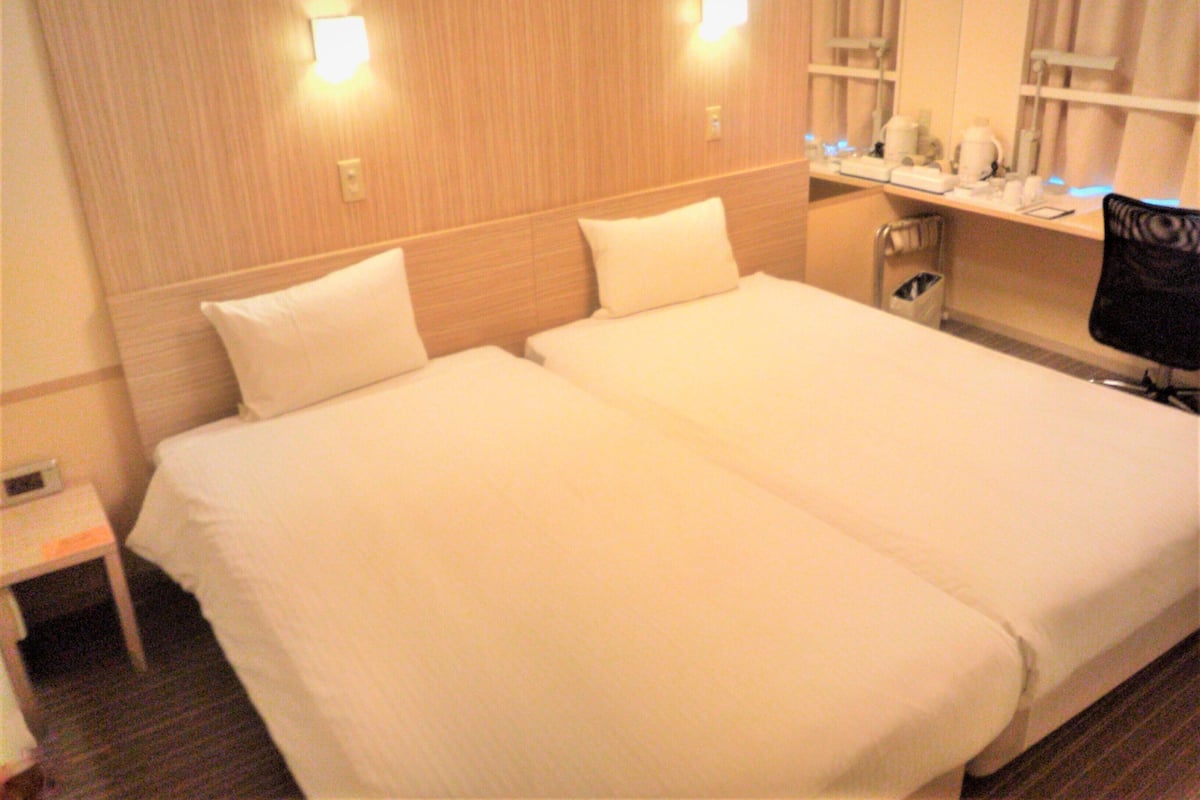 Quarto de hotel com duas camas, uma mesinha de cabeceira, uma cadeira e um espelho.