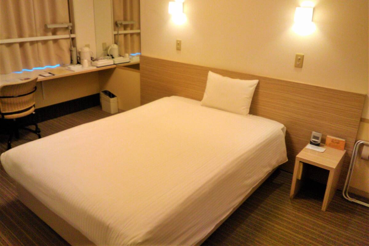 Quarto de hotel com uma cama grande, uma mesa de trabalho e uma cadeira. Há duas luminárias fixadas na parede e uma pequena mesa com uma cadeira.