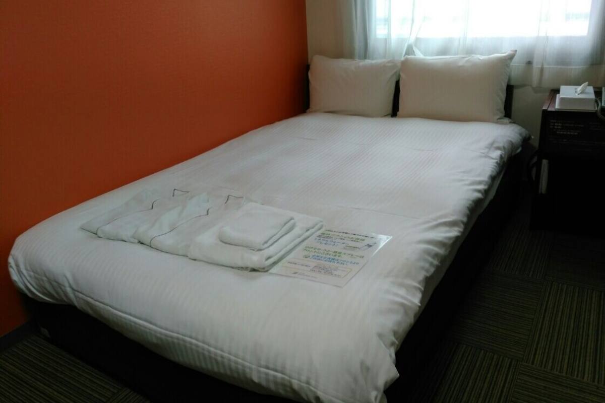 Una cama bien hecha con sábanas blancas y una toalla doblada, una mesita de noche con un pañuelito, y una ventana con cortinas traslúcidas.