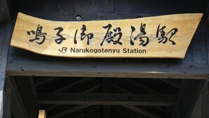 Uma placa de madeira com caracteres japoneses e a inscrição em inglês 'Narukogotenyu Station'.