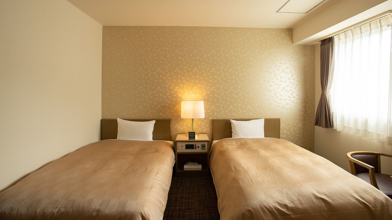A hotel room with two single beds, a bedside table with a lamp, a small chair, and a window with curtains.