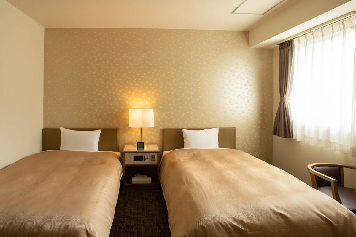 A hotel room with two single beds, a bedside table with a lamp, a small chair, and a window with curtains.