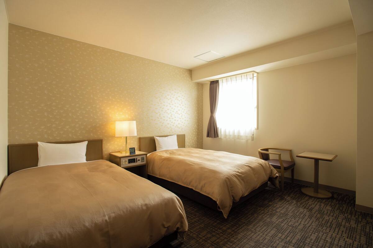 A hotel room with two beds, a nightstand with a lamp, a small table, and a window with curtains.
