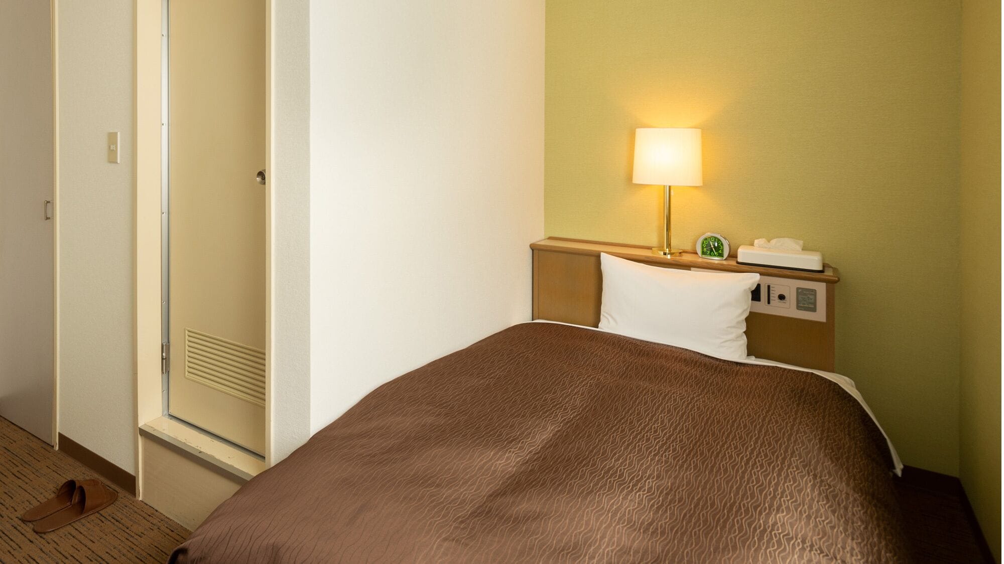 A hotel room with a bed, a nightstand, a lamp, a clock, and a door.
