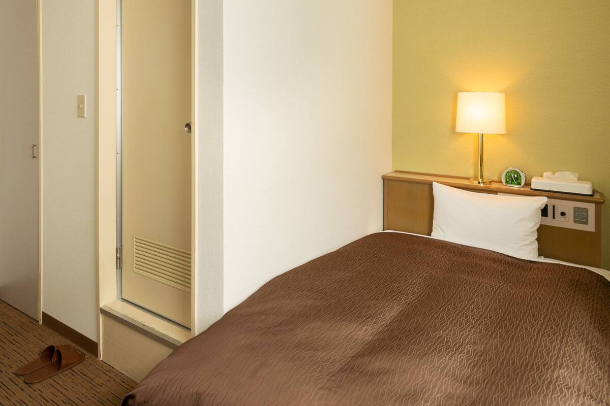 A hotel room with a bed, a nightstand, a lamp, a clock, and a door.