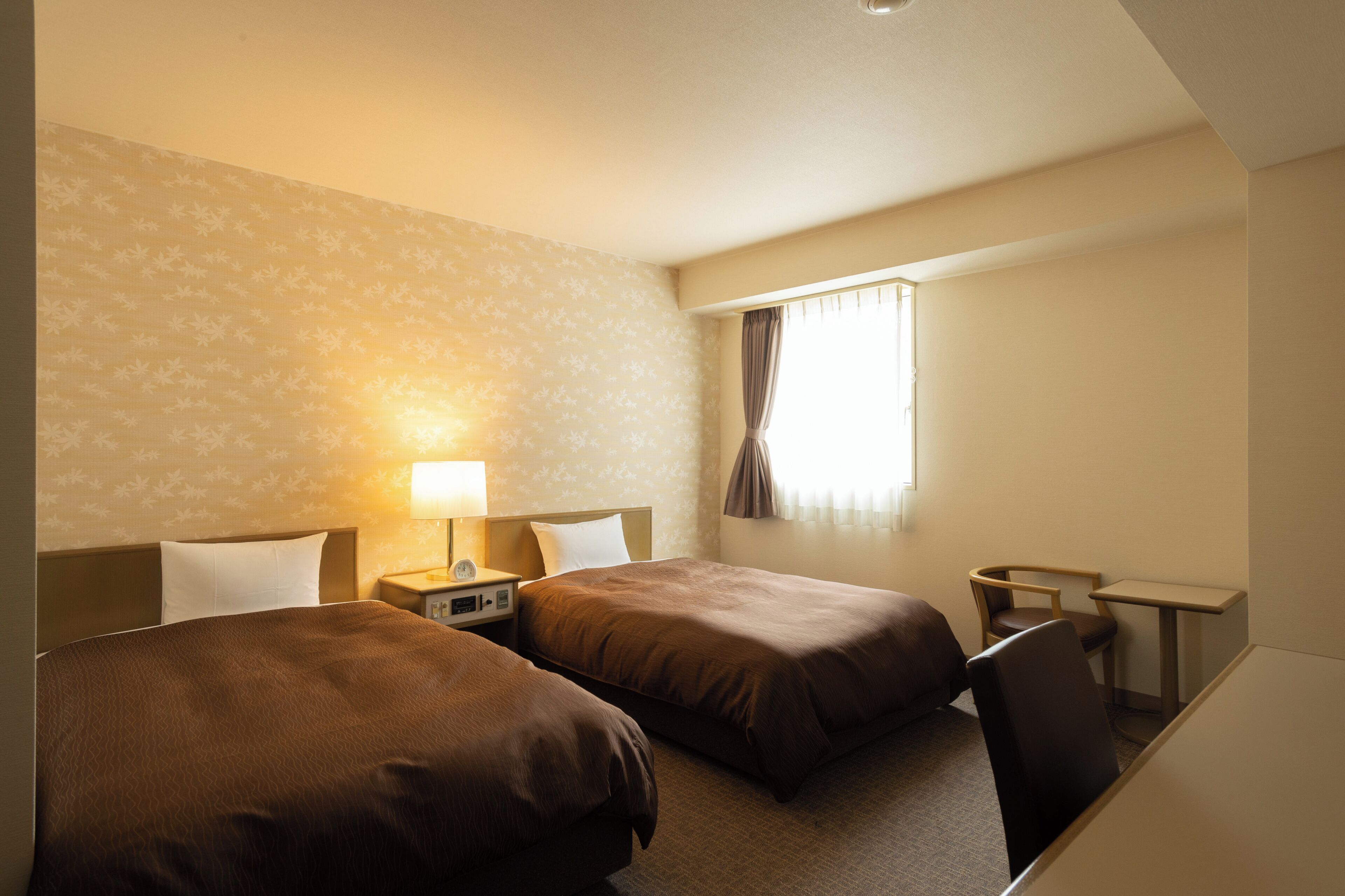 A hotel room with two beds, a desk, a chair, a lamp, and a window with curtains.