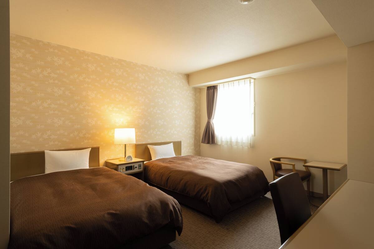 A hotel room with two beds, a desk, a chair, a lamp, and a window with curtains.