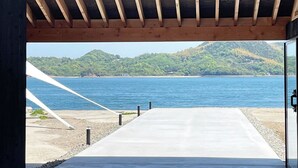 A view through a wooden structure with a concrete platform leading to a body of water, surrounded by greenery and hills.
