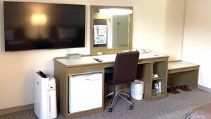 A hotel room with a desk, a television, a mirror, a mini-fridge, a trash bin, and a shoe rack.