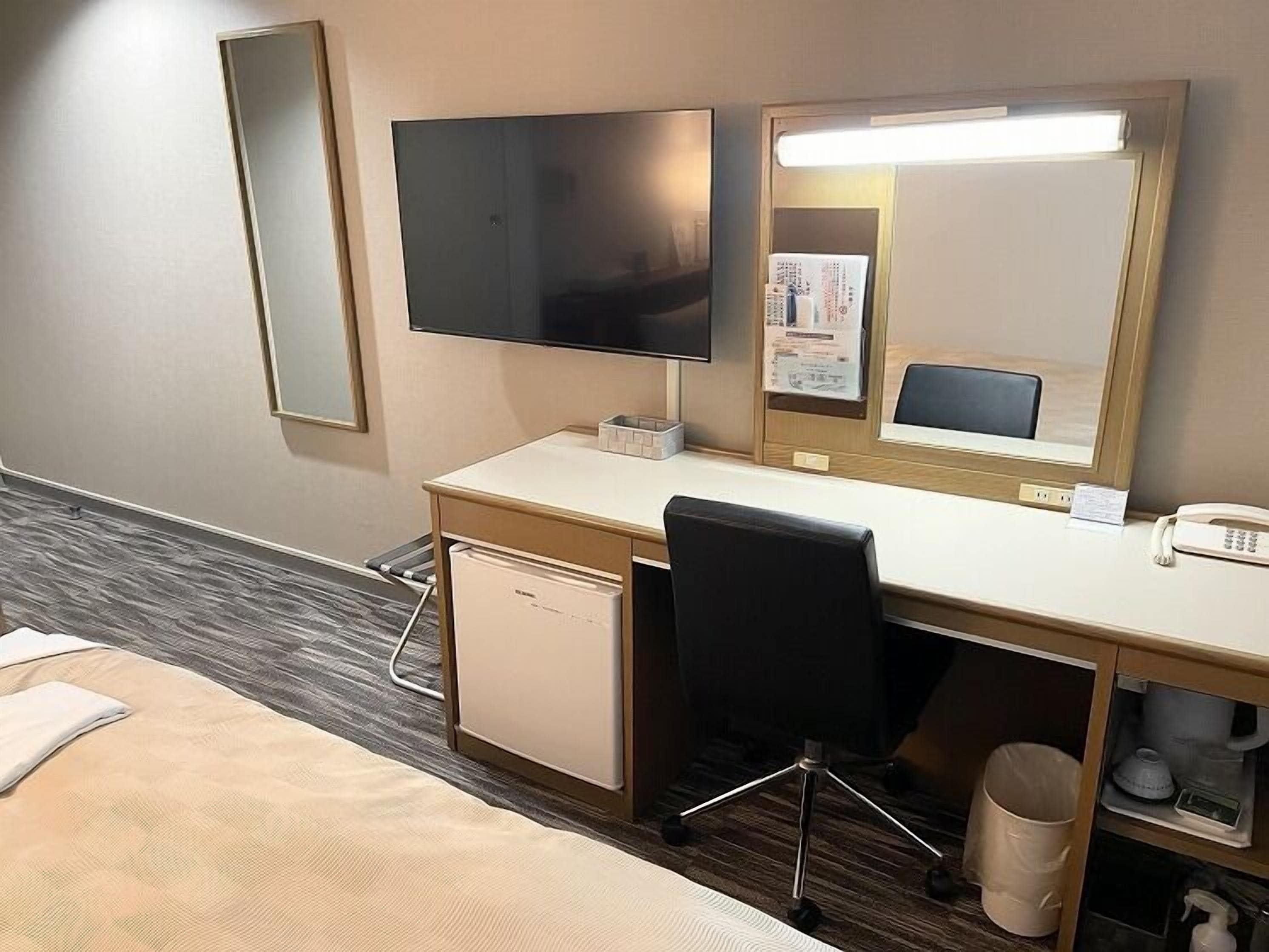 A hotel room with a bed, a desk with a chair, a television, a mirror, a telephone, and a small refrigerator.