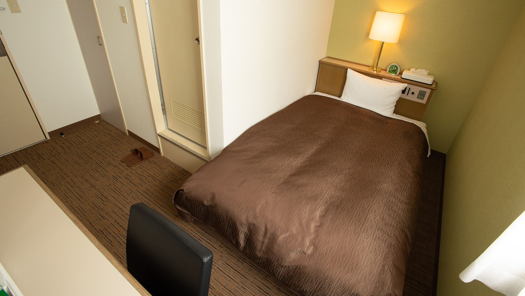 A hotel room with a bed, a desk, a chair, a nightstand with a lamp, and a window.
