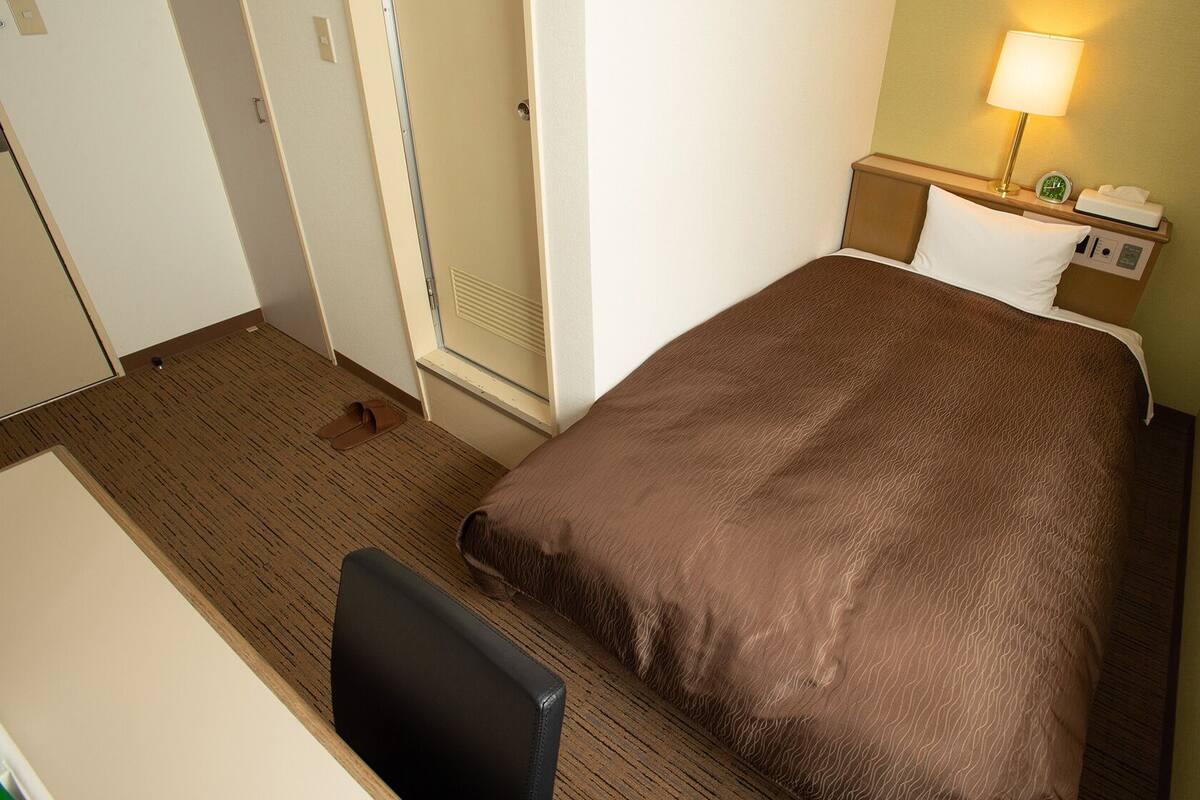 A hotel room with a bed, a desk, a chair, a nightstand with a lamp, and a window.
