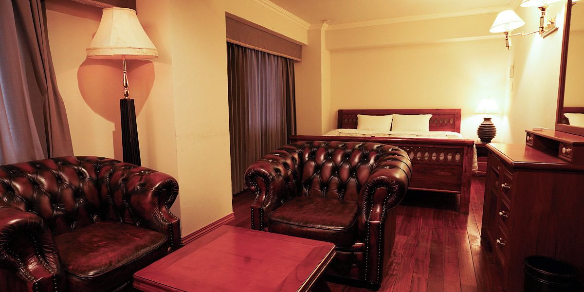 A hotel room with a bed, two leather armchairs, a wooden coffee table, a nightstand, and a lamp.