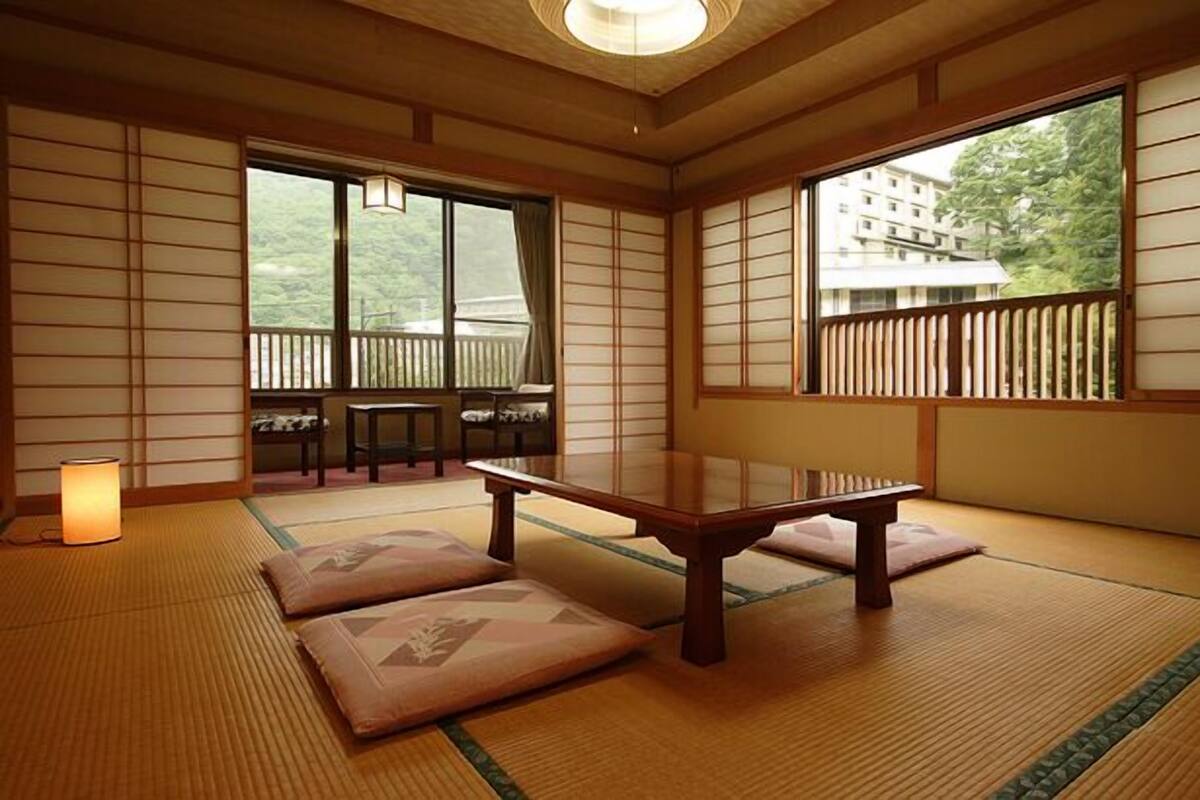 A traditional Japanese room with tatami flooring, a low table, cushions, sliding doors, and a view of greenery.