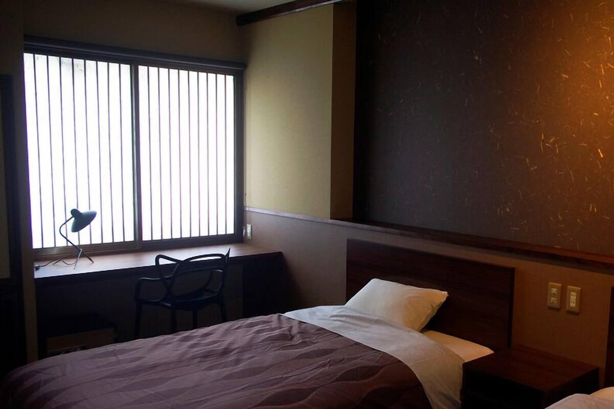 A hotel room with a bed, a desk with a chair, a window with blinds, and a large wall with a textured surface.