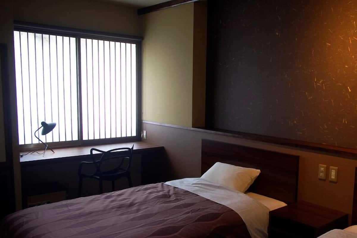 A hotel room with a bed, a desk with a chair, a window with blinds, and a large wall with a textured surface.
