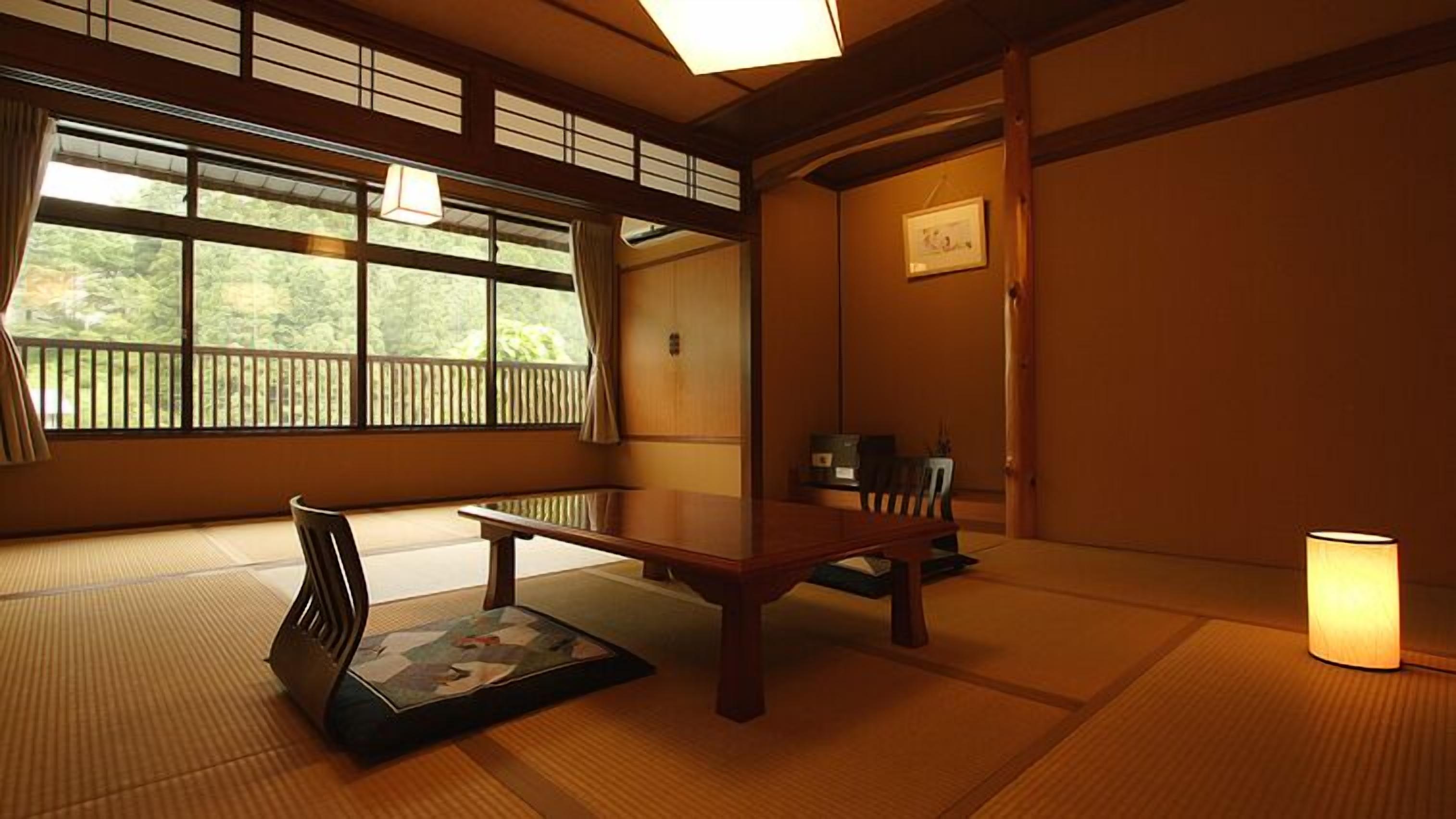 A traditional Japanese room with tatami flooring, a low table, a chair, a hanging lamp, and a view through large windows.