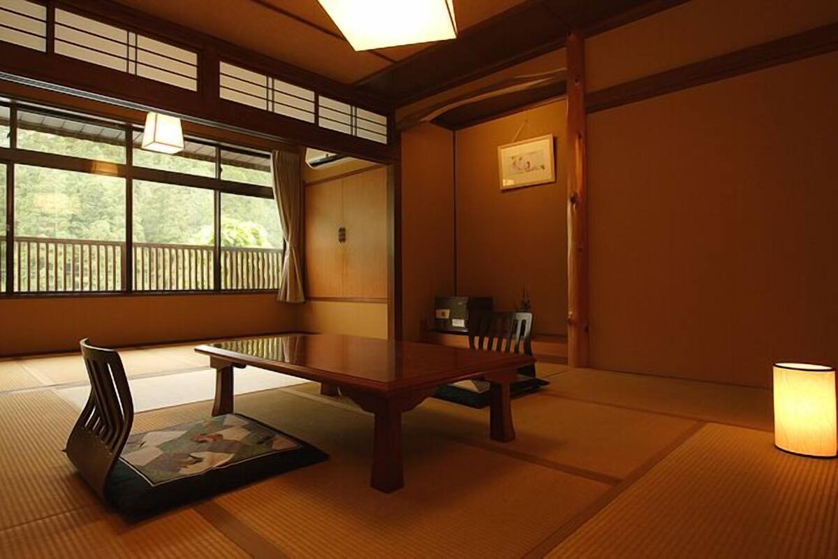 A traditional Japanese room with tatami flooring, a low table, a chair, a hanging lamp, and a view through large windows.