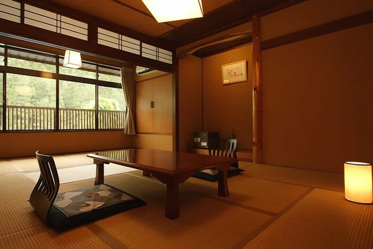A traditional Japanese room with tatami flooring, a low table, a chair, a hanging lamp, and a view through large windows.