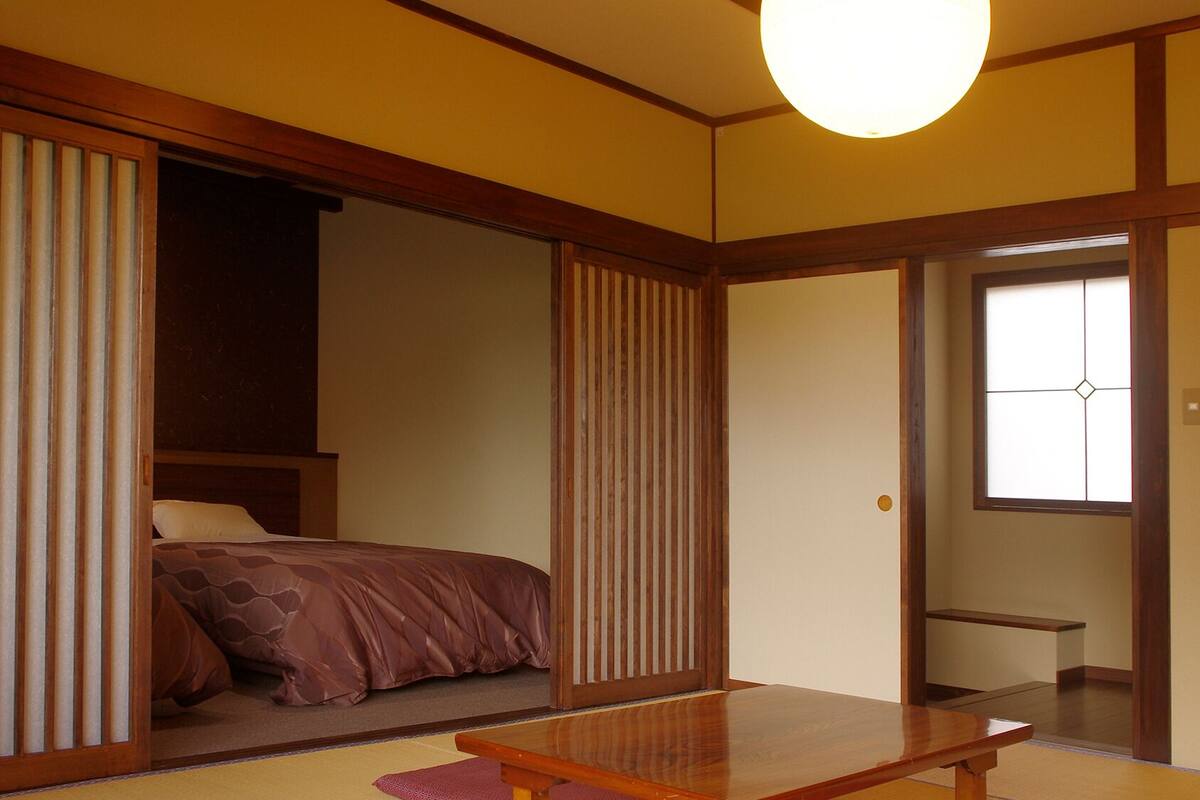 A traditional Japanese room with tatami flooring, a low wooden table, a bed with a brown bedspread, and sliding shoji doors.