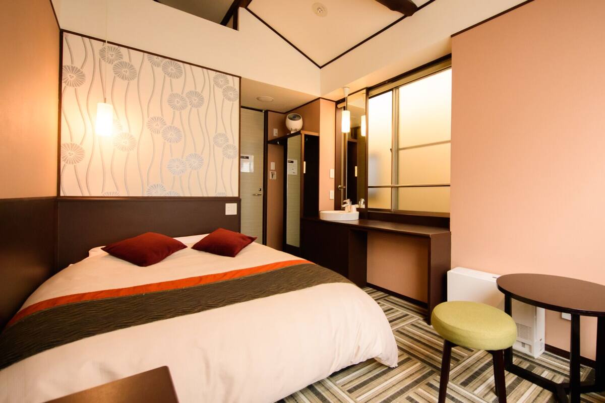 A hotel room with a bed, a small table, a chair, a mirror, and a window.