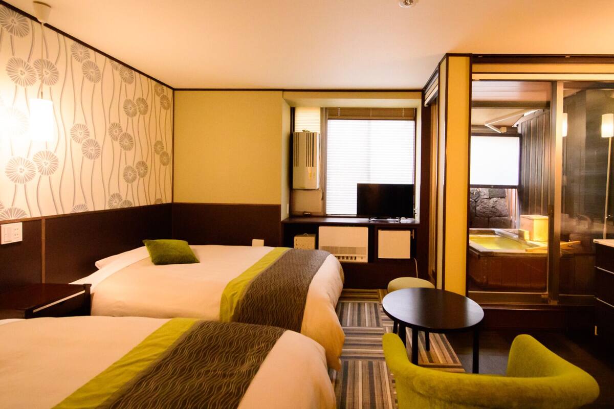 A hotel room with two beds, a small table, a chair, a TV, and a bathroom with a bathtub.