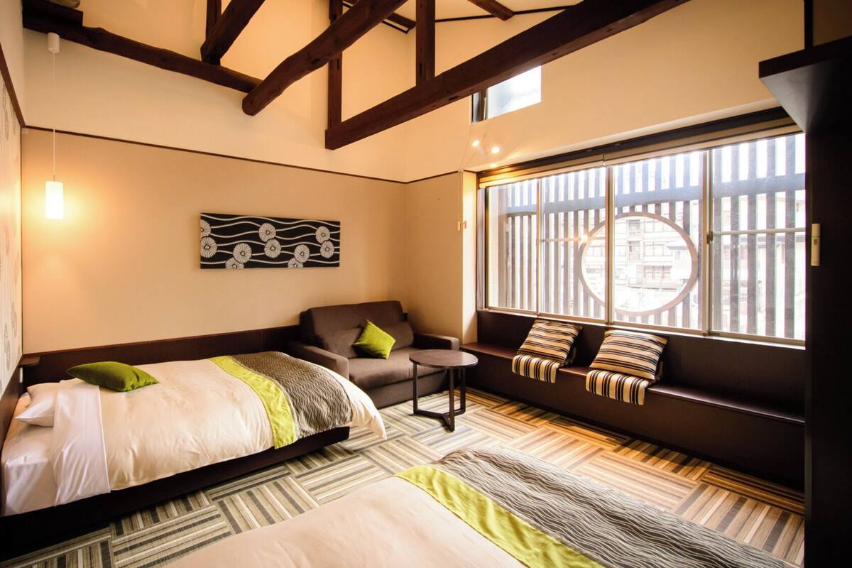 A hotel room with a bed, a sofa, a small table, and a window with a view of buildings.