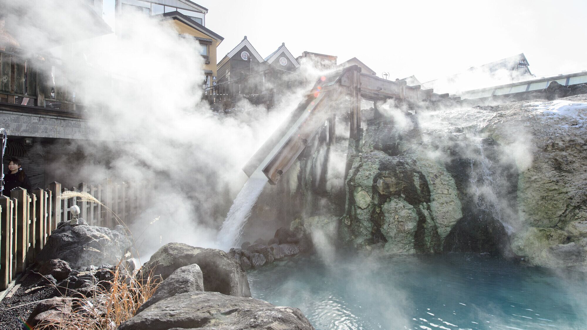 A hot spring area with a wooden walkway, rocks, and a building in the background.