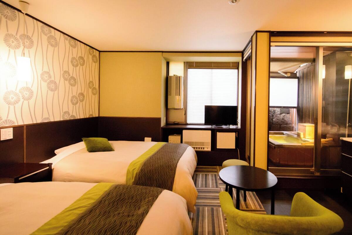 A hotel room with two beds, a small table, a chair, a TV, and a bathroom with a bathtub.